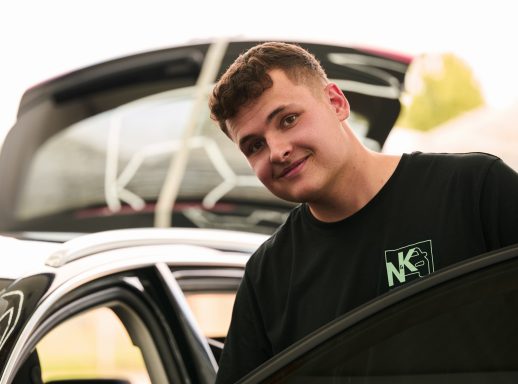 Junger Mann steht lächelnd neben einem Auto mit offenem Kofferraum.
