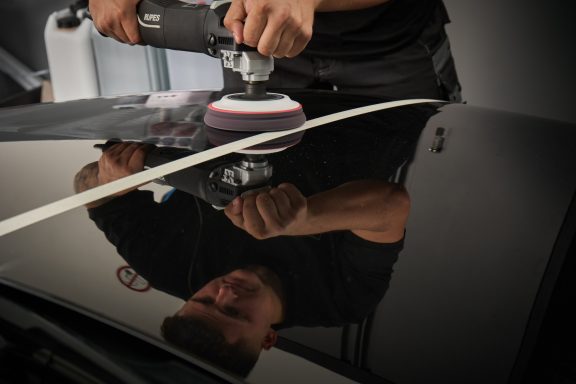 Eine Person poliert mit einer Maschine eine schwarze Autolackoberfläche.