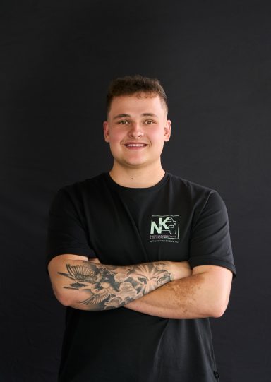 Junger Mann mit kurzen Haaren, lächelnd, in schwarzem T-Shirt, vor schwarzem Hintergrund.